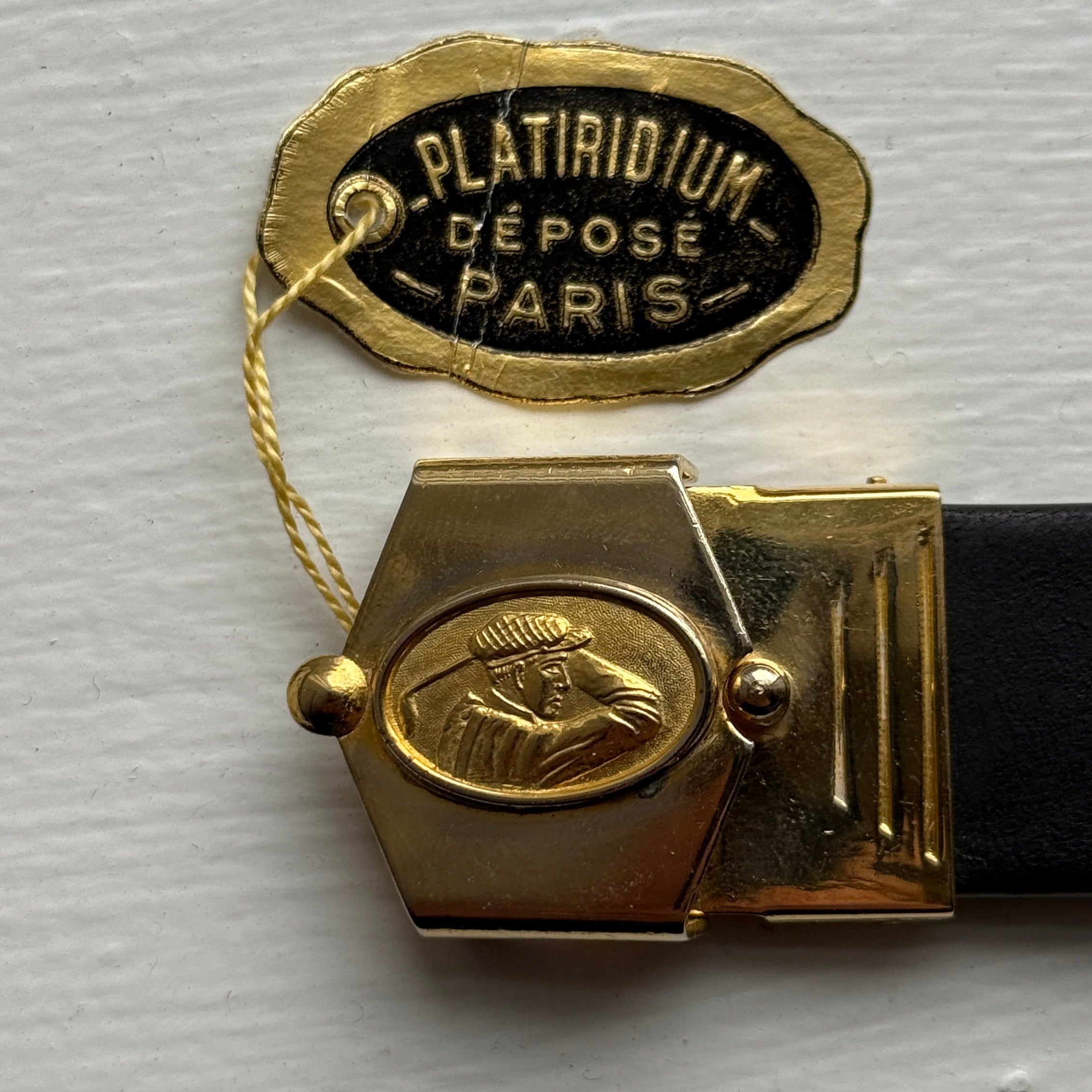 Gold belt buckle with 'Platinum Depose Paris' label on a white background
